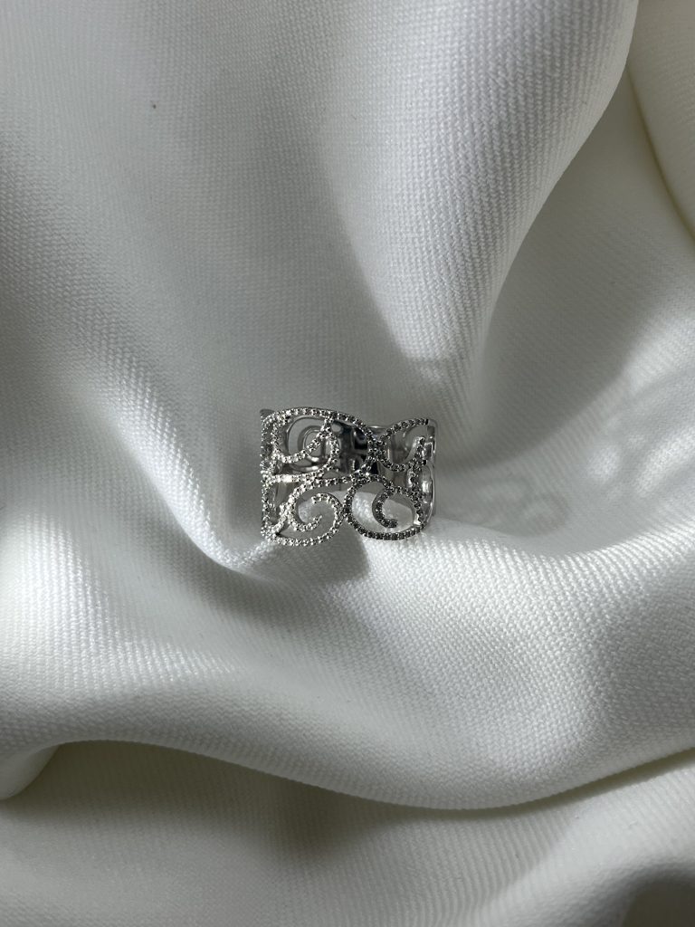 Bague large ajourée en or blanc 18 carats, motif d'arabesques serties de diamants, posée sur un tissu blanc Si vous avez d'autres photos du même modèle, quelques variantes pour éviter les doublons : Vue de profil : Bague ajourée or blanc et diamants vue de profil, montrant la largeur de l'anneau Portée : Bague large arabesques or blanc et diamants portée à la main Détail : Détail du serti des diamants sur les volutes de la bague ajourée or blanc
