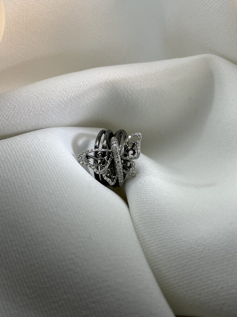Bague papillon en or rhodié noir et blanc, ailes ajourées serties de diamants, sur un anneau à plusieurs rangs, posée sur un drapé de tissu blanc.